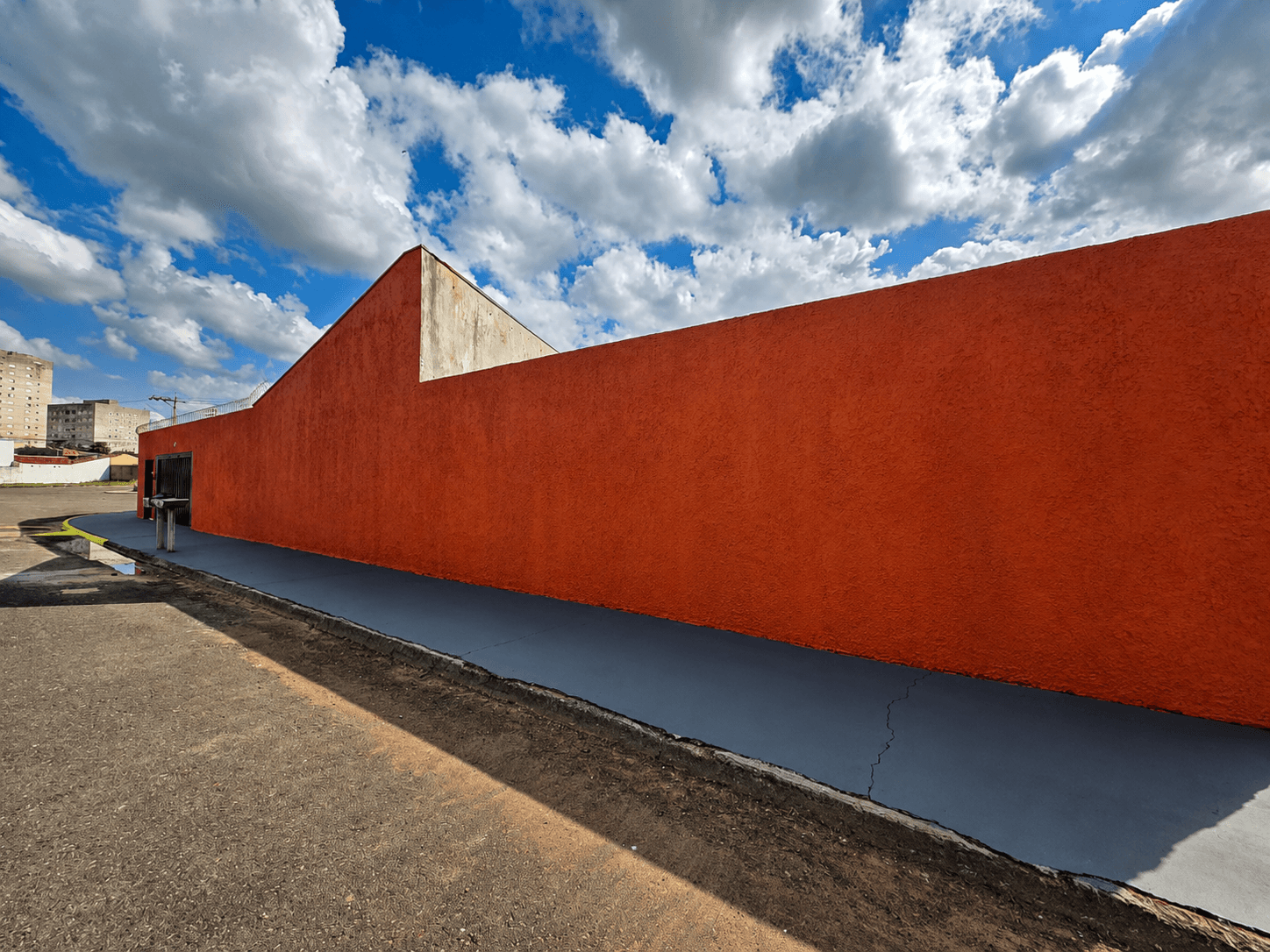 Muro lateral laranja da Vila Agostinis, com arame farpado no topo, sob céu azul com nuvens.