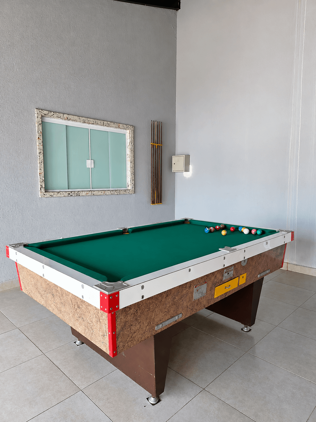 Mesa de sinuca da Vila Agostinis com suporte de tacos na parede e janela ao fundo.